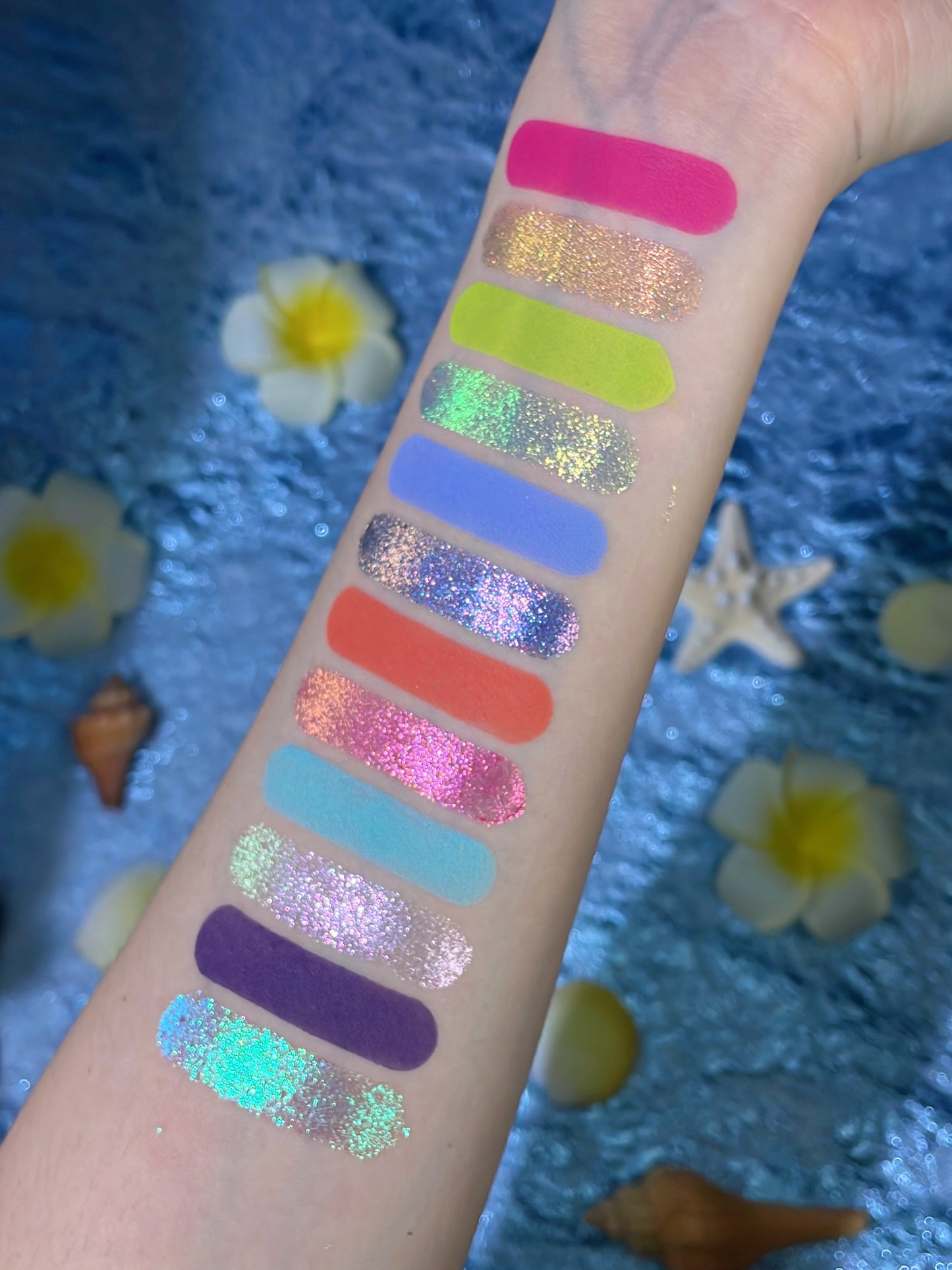 Tropical Mirage arm swatches from a second angle, the same 12 shades under different light against a blue backdrop with plumeria and shells