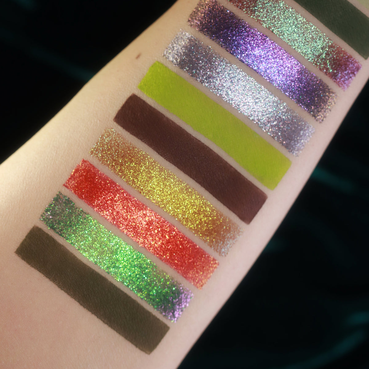 Close-up of arm swatches on light skin: purple and silver sparkles, lime and plum mattes, gold, red and green sparkles, olive matte