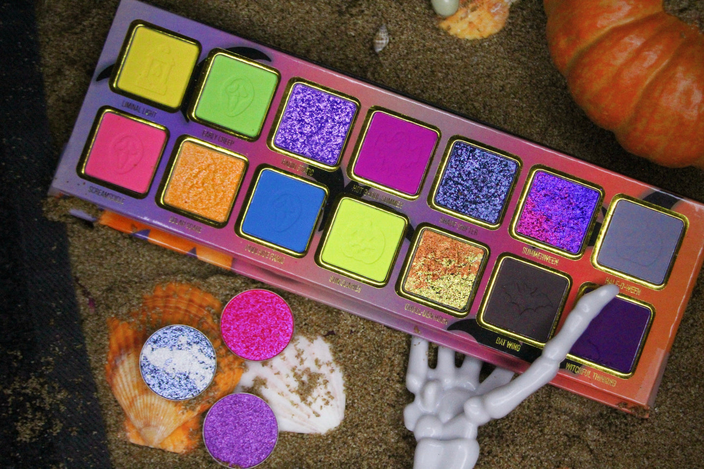Summerween palette in sand with seashells, a small pumpkin, and a skeleton hand, with three loose sparkle pans set in the shells