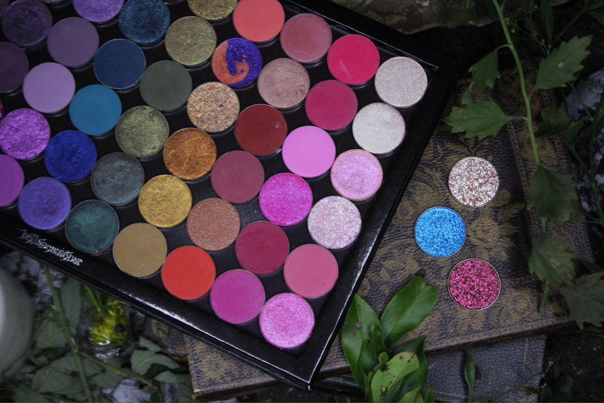 Display shot of a magnetic palette filled with matte and shimmer singles, three loose pans beside it on dark foliage