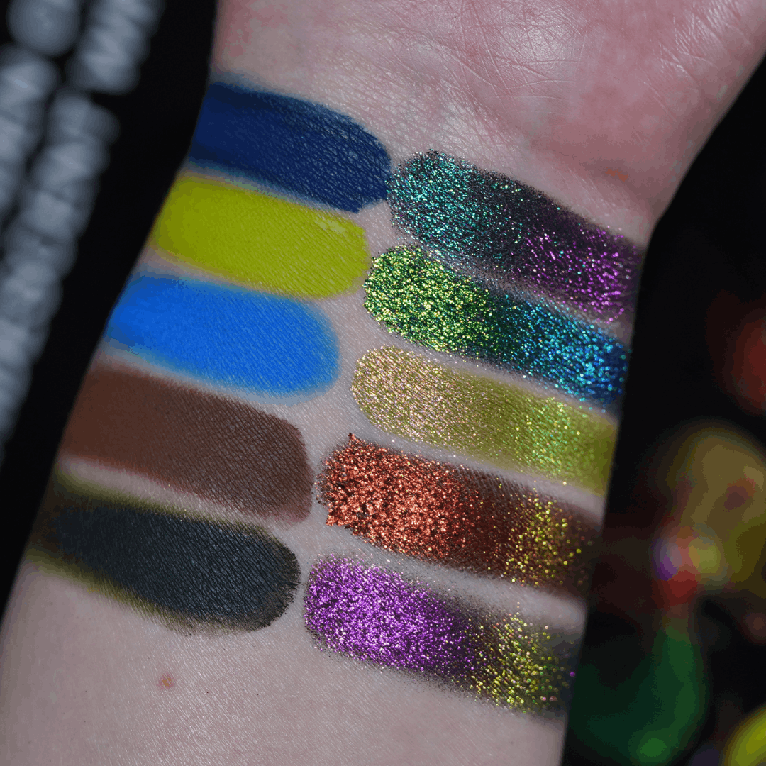 All ten shades swatched as oval patches on a fair wrist, matte yellows browns and black beside shimmer greens, copper and magenta multichromes.