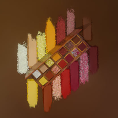 Pandemonium palette tilted over powder swatch streaks on a brown backdrop, whites, yellows, oranges, reds, pinks and sparkles laid out behind it