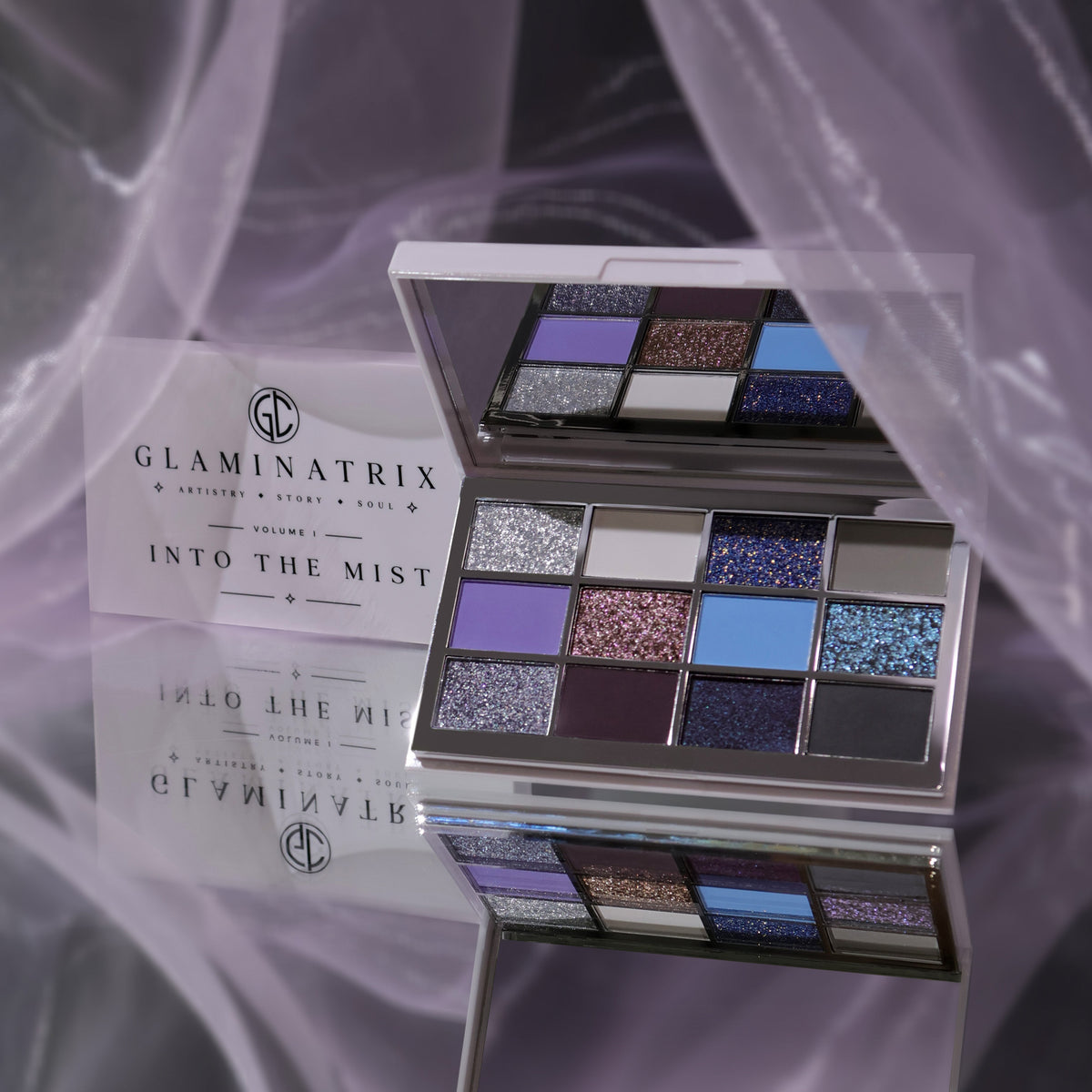 Glaminatrix Into the Mist eyeshadow palette open with its mirror, 12 silver, lavender, blue, plum and grey shades, lilac sheer fabric behind