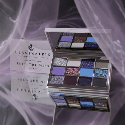 Glaminatrix Into the Mist eyeshadow palette open with its mirror, 12 silver, lavender, blue, plum and grey shades, lilac sheer fabric behind