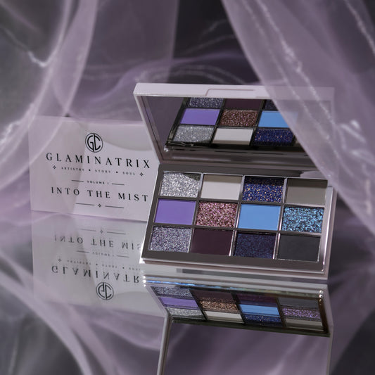 Glaminatrix Into the Mist eyeshadow palette open with its mirror, 12 silver, lavender, blue, plum and grey shades, lilac sheer fabric behind