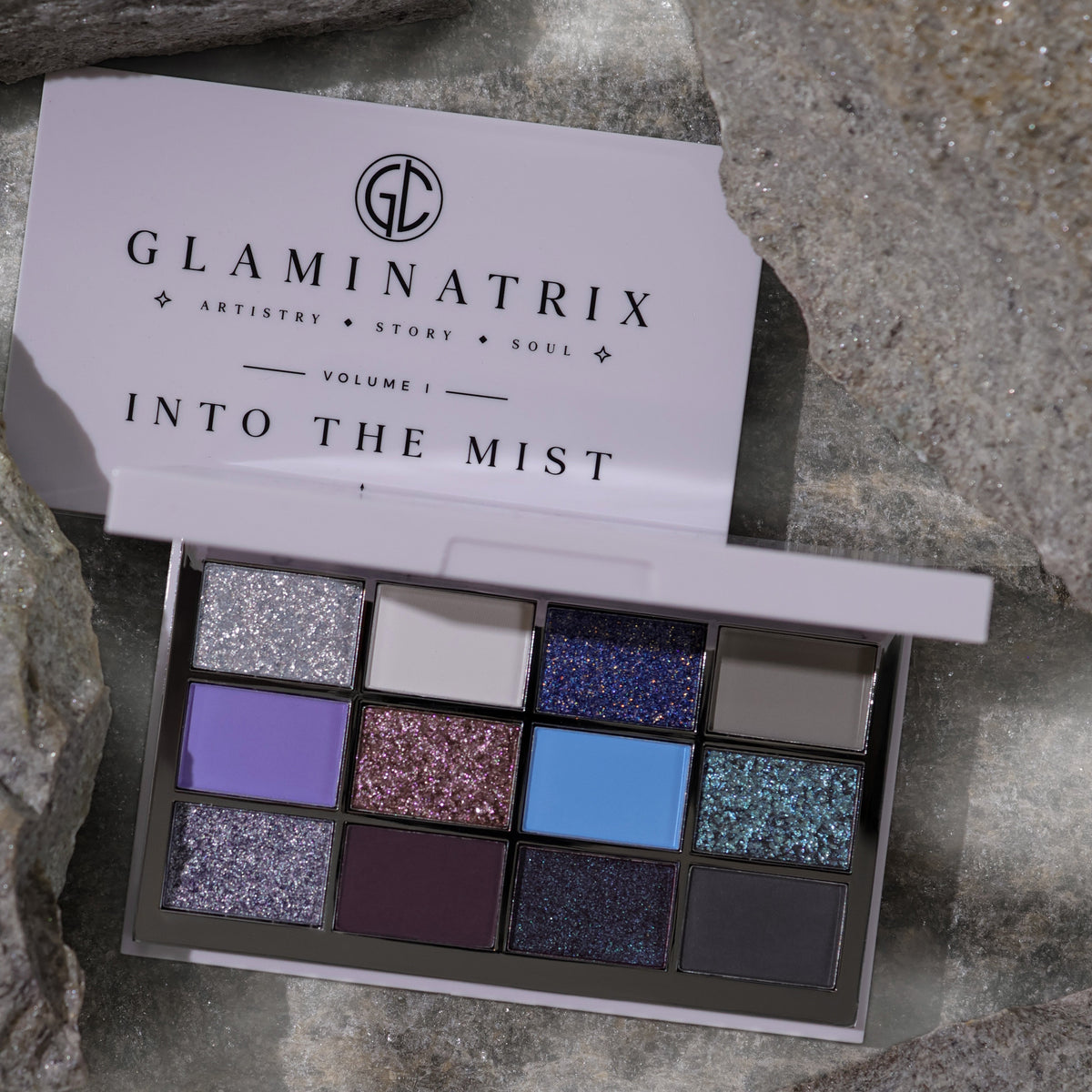 Into the Mist palette open on grey stone beneath its white Volume I lid, three rows of sparkles and mattes in silver, violet, blue and plum