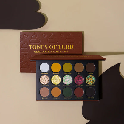 Glaminatrix Cosmetics Tones of Turd eyeshadow palette open, brown poop-embossed lid above 15 pans: warm mattes, green-pink multichromes, and a metallic shimmer