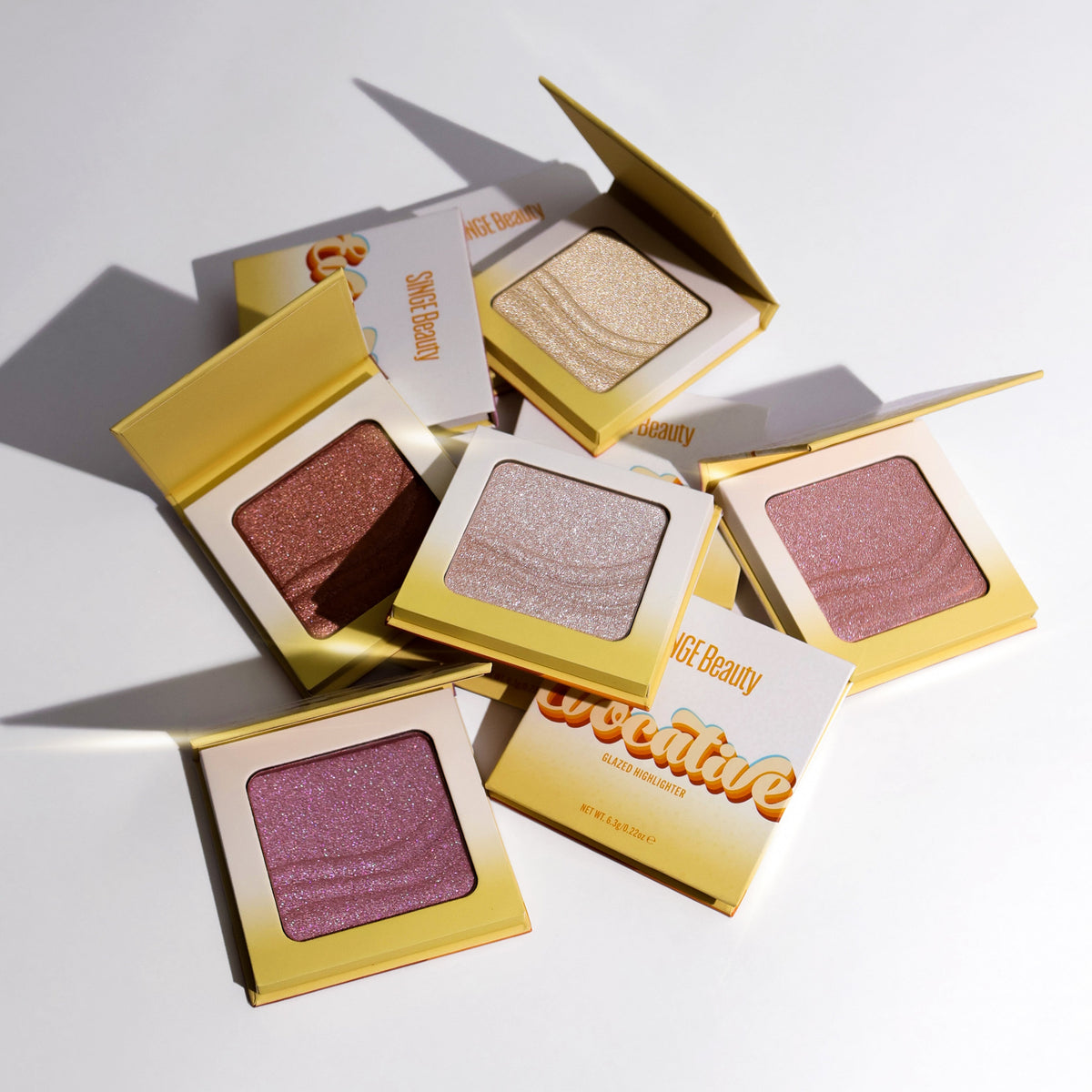 All five Evocative glazed highlighters open and closed in yellow and white compacts, scattered on white with two retro-lettered boxes