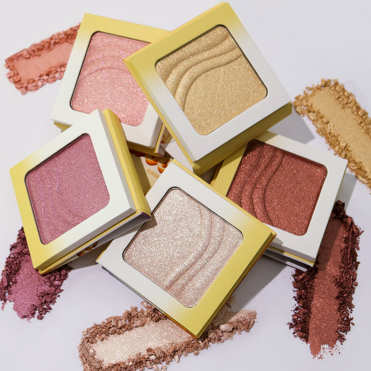 The five Evocative glazed highlighter compacts stacked over crushed swatches of each shade on a pale lilac background