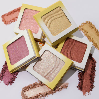 The five Evocative glazed highlighter compacts stacked over crushed swatches of each shade on a pale lilac background