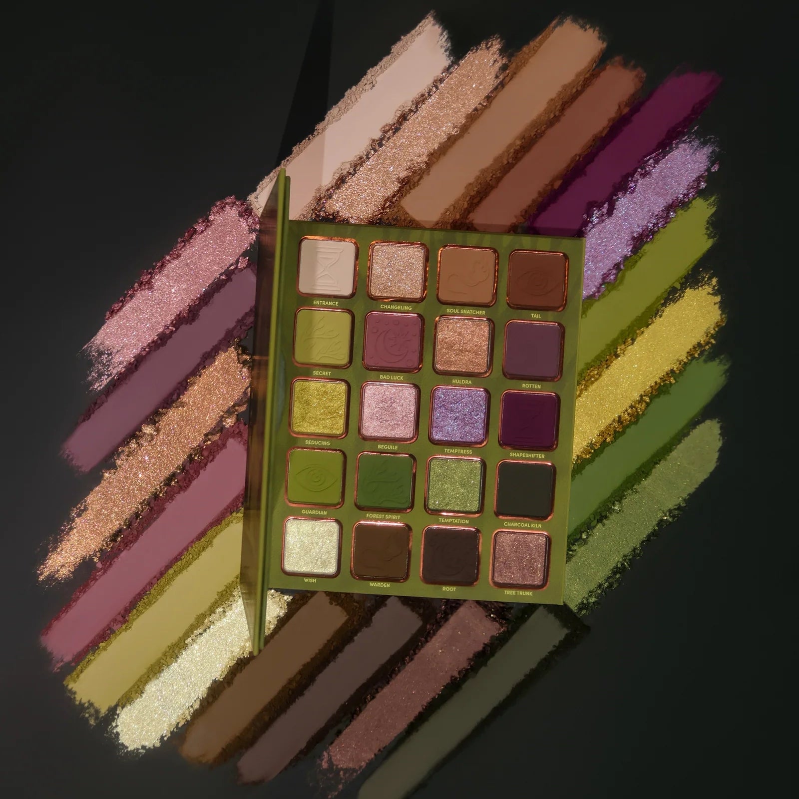 The Huldra palette open top-down against streaks of crushed pigment in greens, mauves, golds, and plums, 20 labelled pans