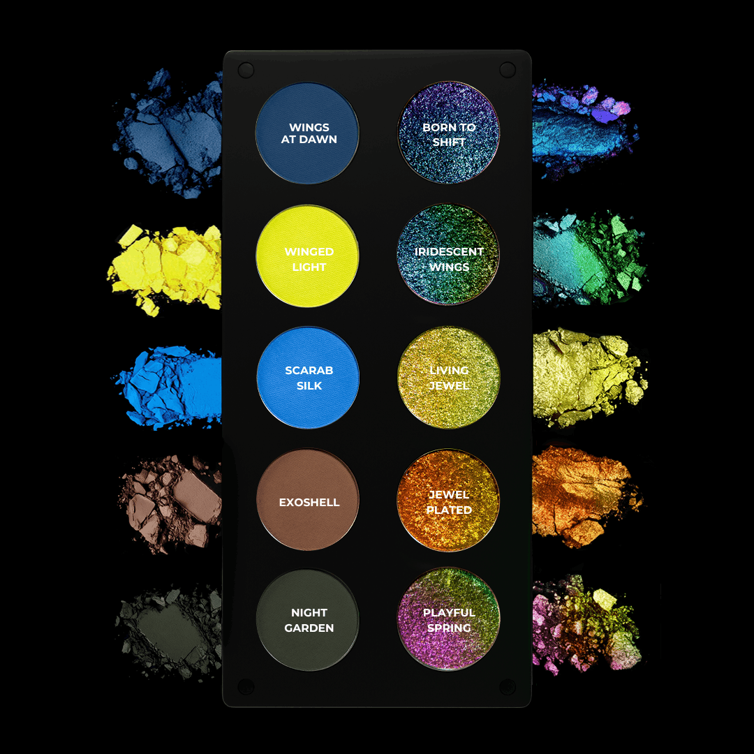 Open palette overhead, ten pans across two rows with shade names printed on each: jewel multichromes and garden shimmers, loose powder scattered around the border.