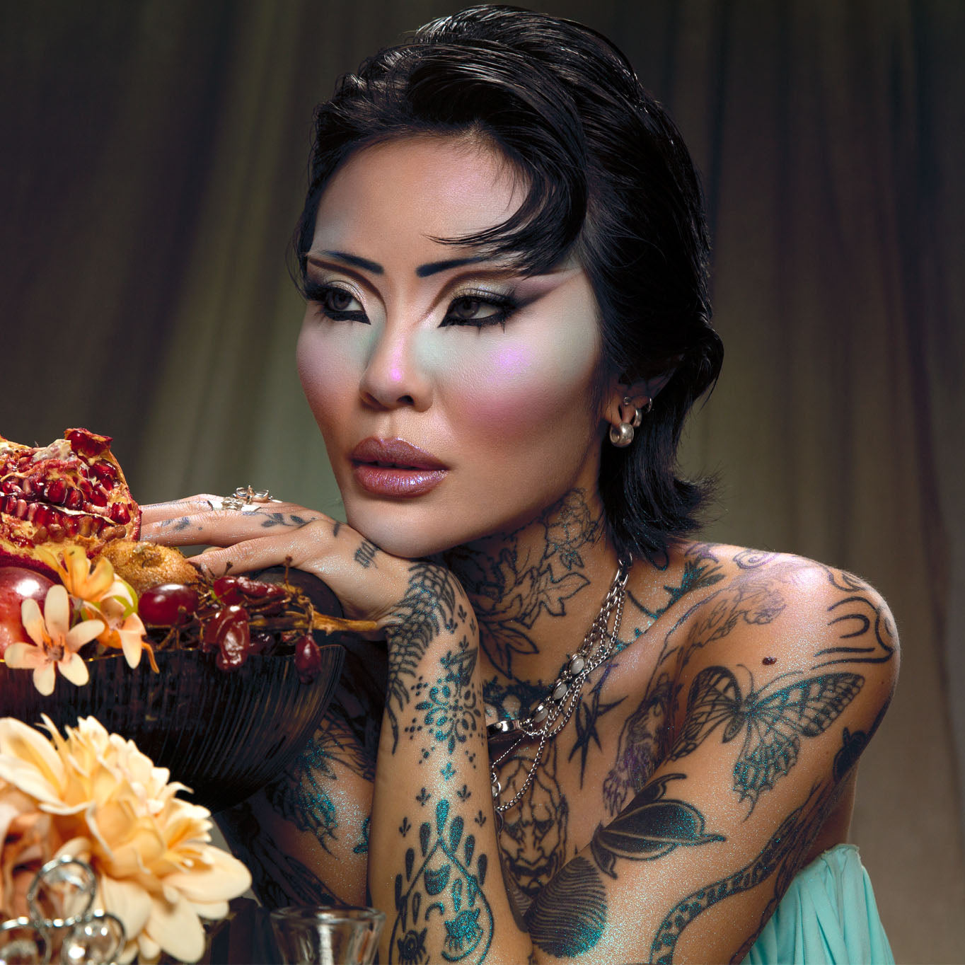 A model with graphic black and pale carved eye makeup and a glossy nude lip, resting on a fruit bowl of pomegranate and grapes, in teal fabric