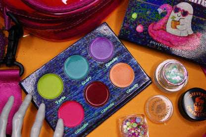 Flat lay on orange: the open palette with a pink bag, loose pigment pots, a pot of gems and the brand's packaging.