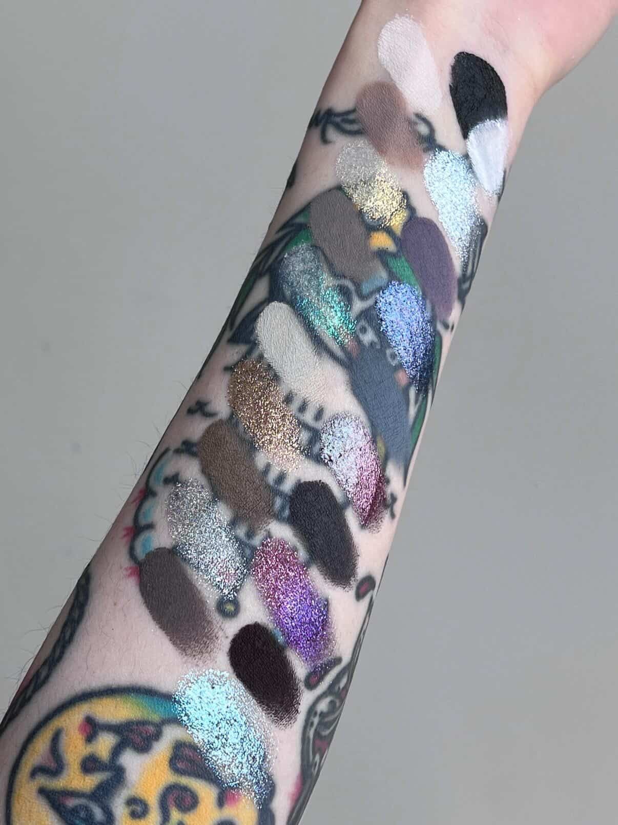 BellaBeauteBar Yin & Yang x Freya86_ff palette full shade swatches on a tattooed arm in natural light — glitters, mattes, and duochromes in grey, purple, brown, and blue