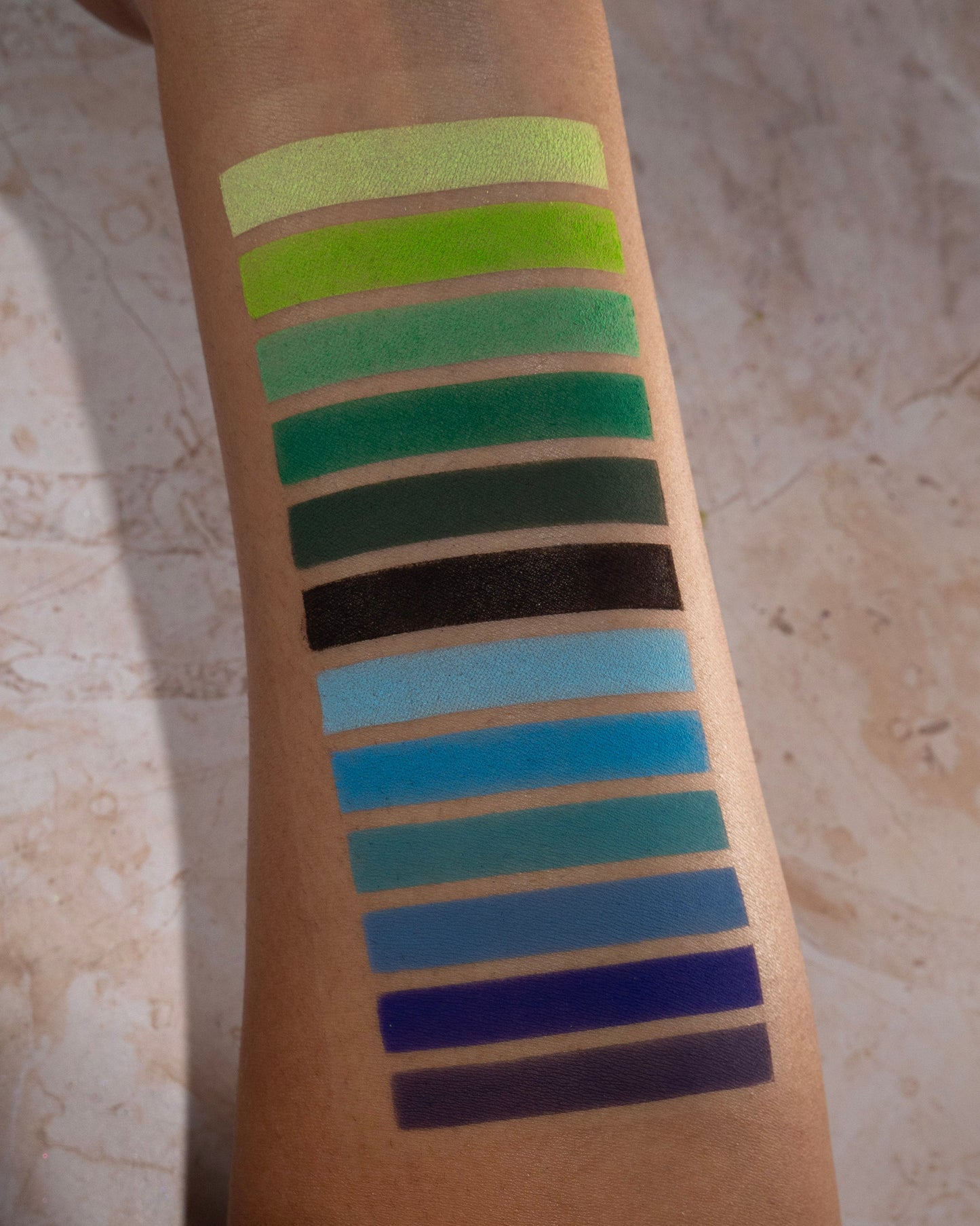 Green to blue shades swatched down a forearm on tan skin, from chartreuse and grass green through teal, black, and indigo