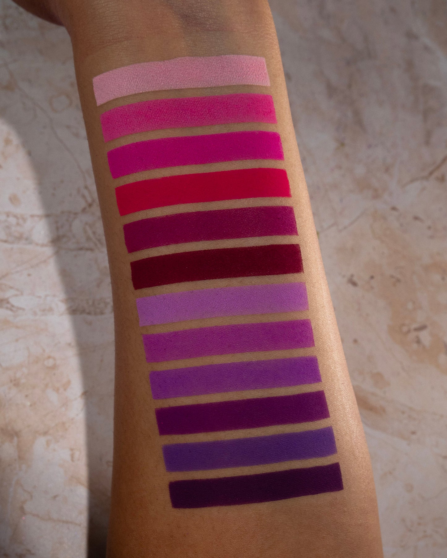 Pink to purple shades swatched down a forearm on tan skin, from soft pink and magenta through red, berry, lilac, and deep violet