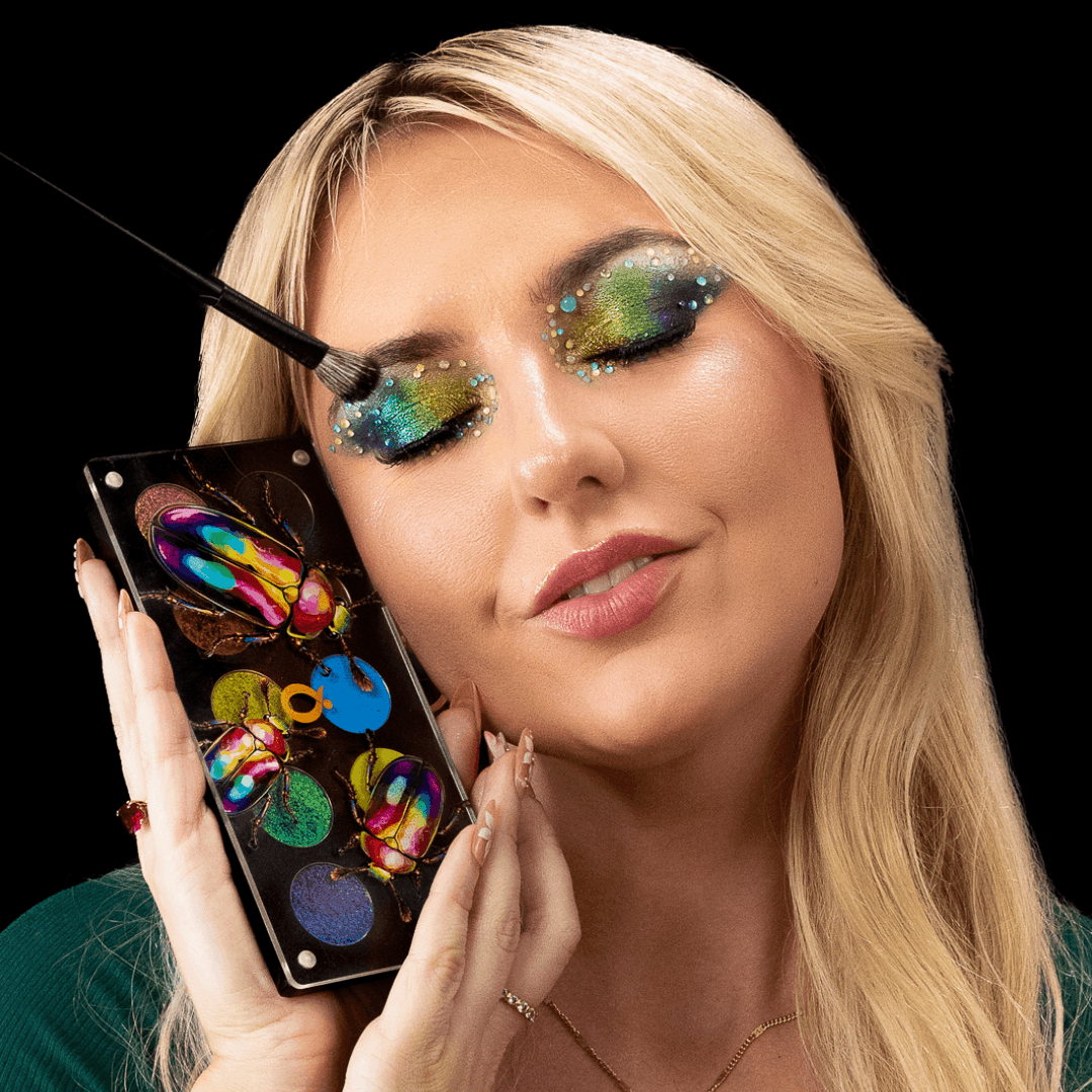 Fair skin blonde model holding the open palette by her cheek and applying a green and blue shimmer eye look with a brush.
