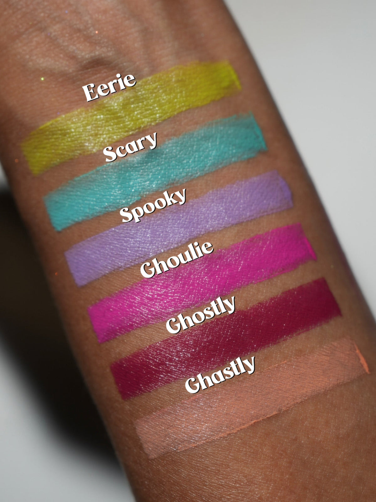 Close-up arm swatches on deep skin in wide bands, labelled Eerie, Scary, Spooky, Ghoulie, Ghostie and Ghastly.