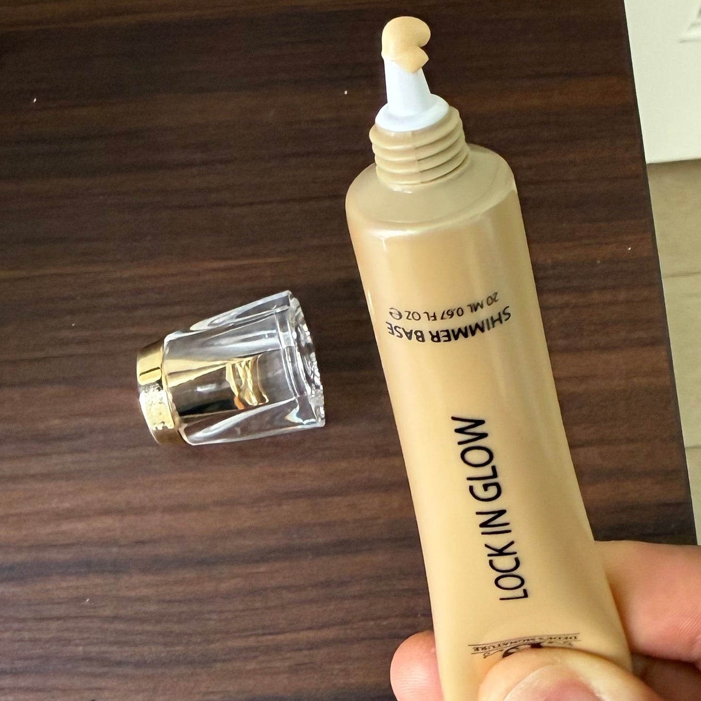 Gold shimmer base tube with the clear gold-rimmed cap removed, pale gold product on the applicator tip, dark wood surface