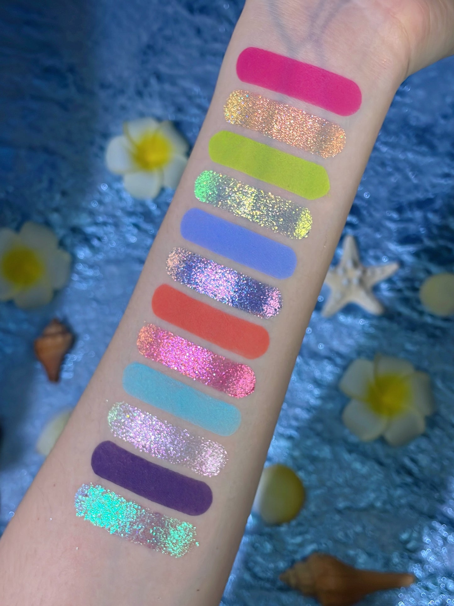 Tropical Mirage arm swatches: 12 shades in one column, mattes beside multichrome sparkles, on a blue backdrop with plumeria, a starfish and shells