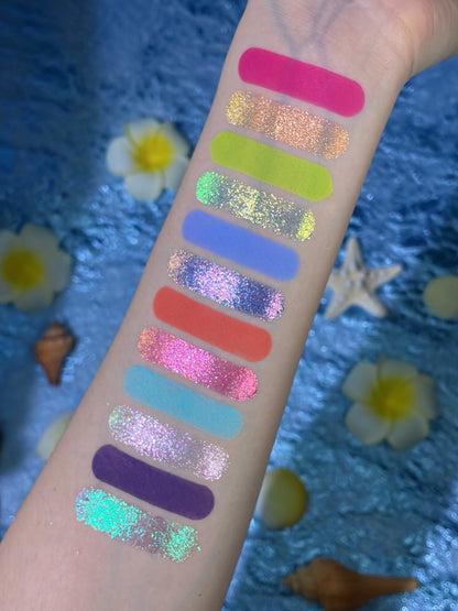 Tropical Mirage arm swatches: 12 shades in one column, mattes beside multichrome sparkles, on a blue backdrop with plumeria, a starfish and shells