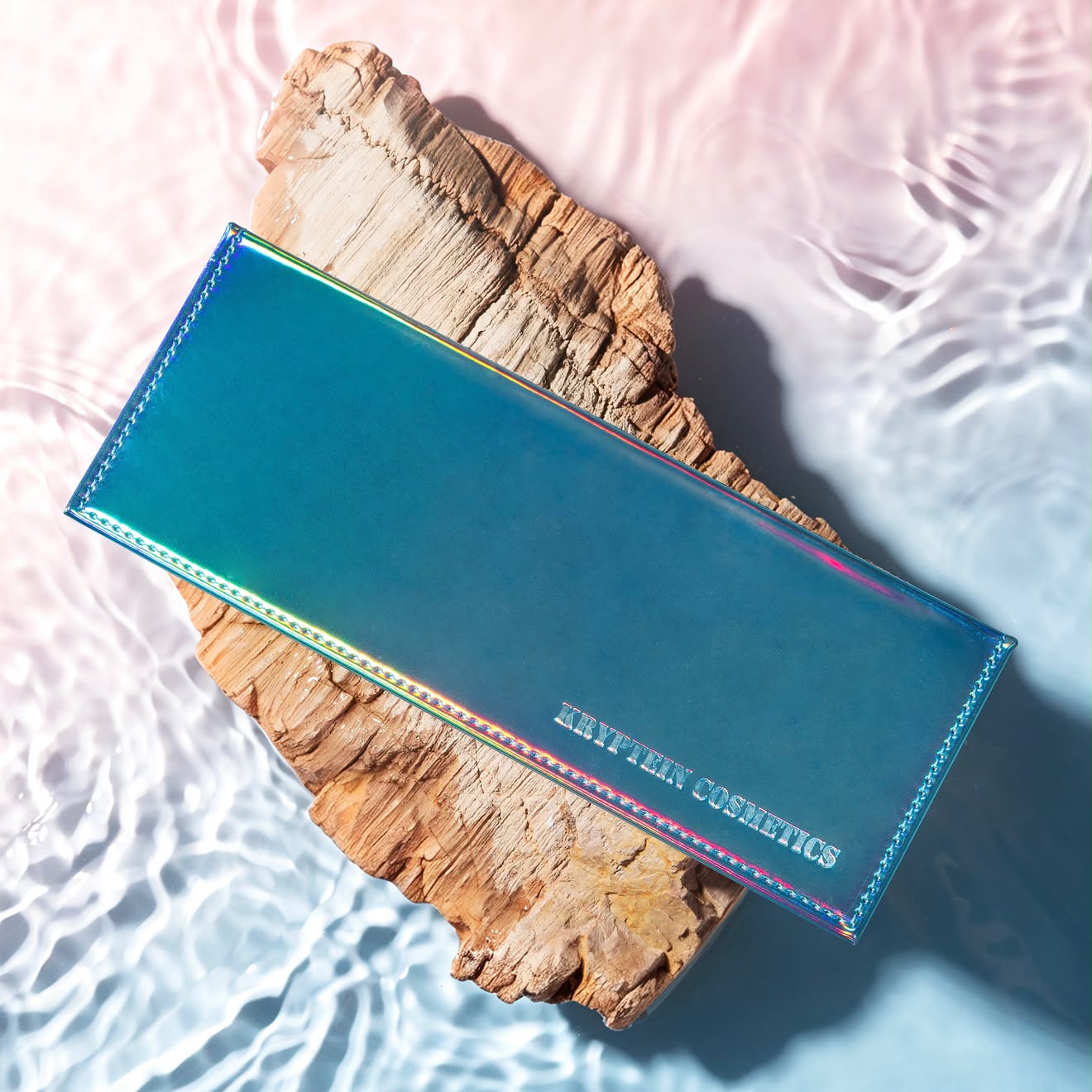 Tropical Mirage palette closed, iridescent teal cover shifting to pink and gold with embossed Kryptein Cosmetics logo, on driftwood in shallow water