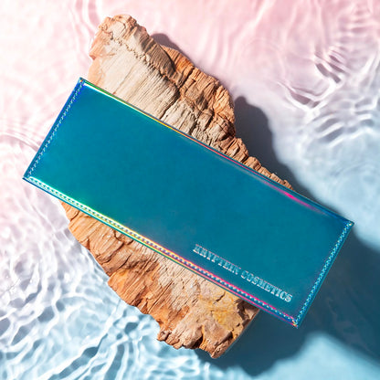 Tropical Mirage palette closed, iridescent teal cover shifting to pink and gold with embossed Kryptein Cosmetics logo, on driftwood in shallow water