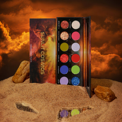 Terra Moons Sunlit Cosmos pressed pigment palette open on sand under an orange sky, showing 12 pans: reds, golds, olive, lime, blues, pink and pale iridescents