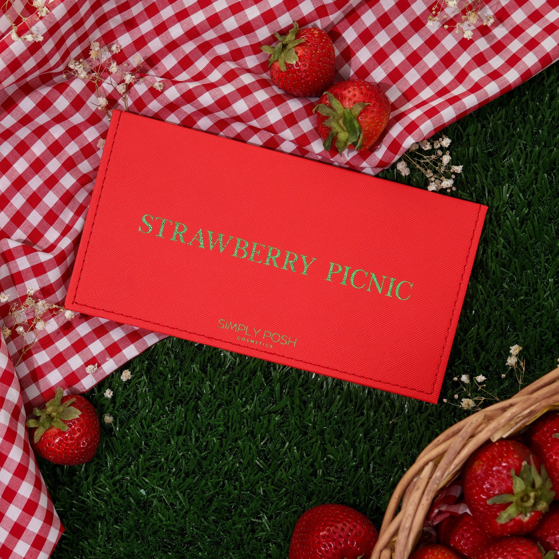 Strawberry Picnic palette closed, red faux-leather cover with gold lettering on red gingham and grass beside a basket of strawberries