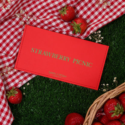 Strawberry Picnic palette closed, red faux-leather cover with gold lettering on red gingham and grass beside a basket of strawberries