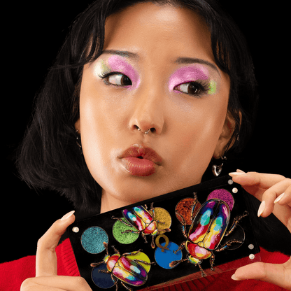 Medium skin tone model holding the open palette under her chin, wearing a hot pink and green shimmer eye look on a red backdrop.