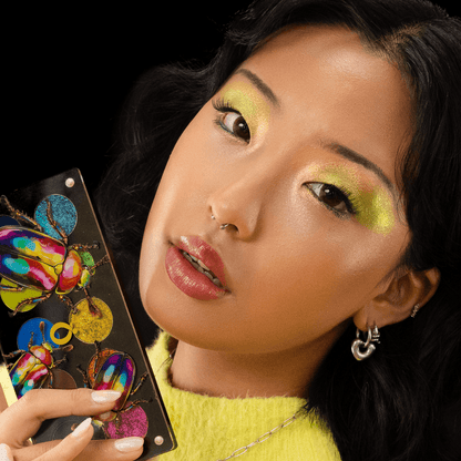 Medium skin tone model holding the open palette by her cheek, wearing a chartreuse yellow shimmer eye look with a fuzzy yellow sweater.