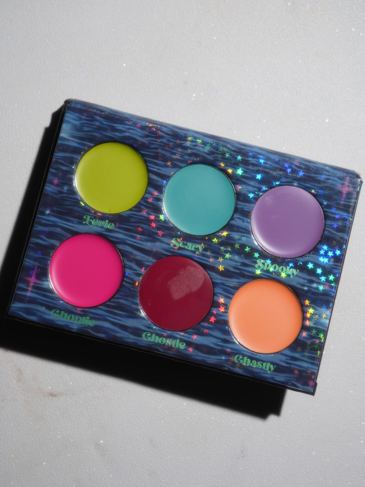 The open palette angled in daylight, six cream pans on a blue water-print tray, each labelled with its shade name.