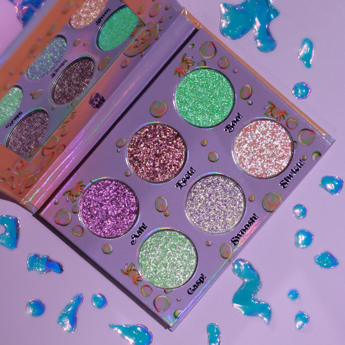 Two open Boodrops palettes overlapping on a lilac surface scattered with blue jelly droplets, the six labelled pans angled to the camera.
