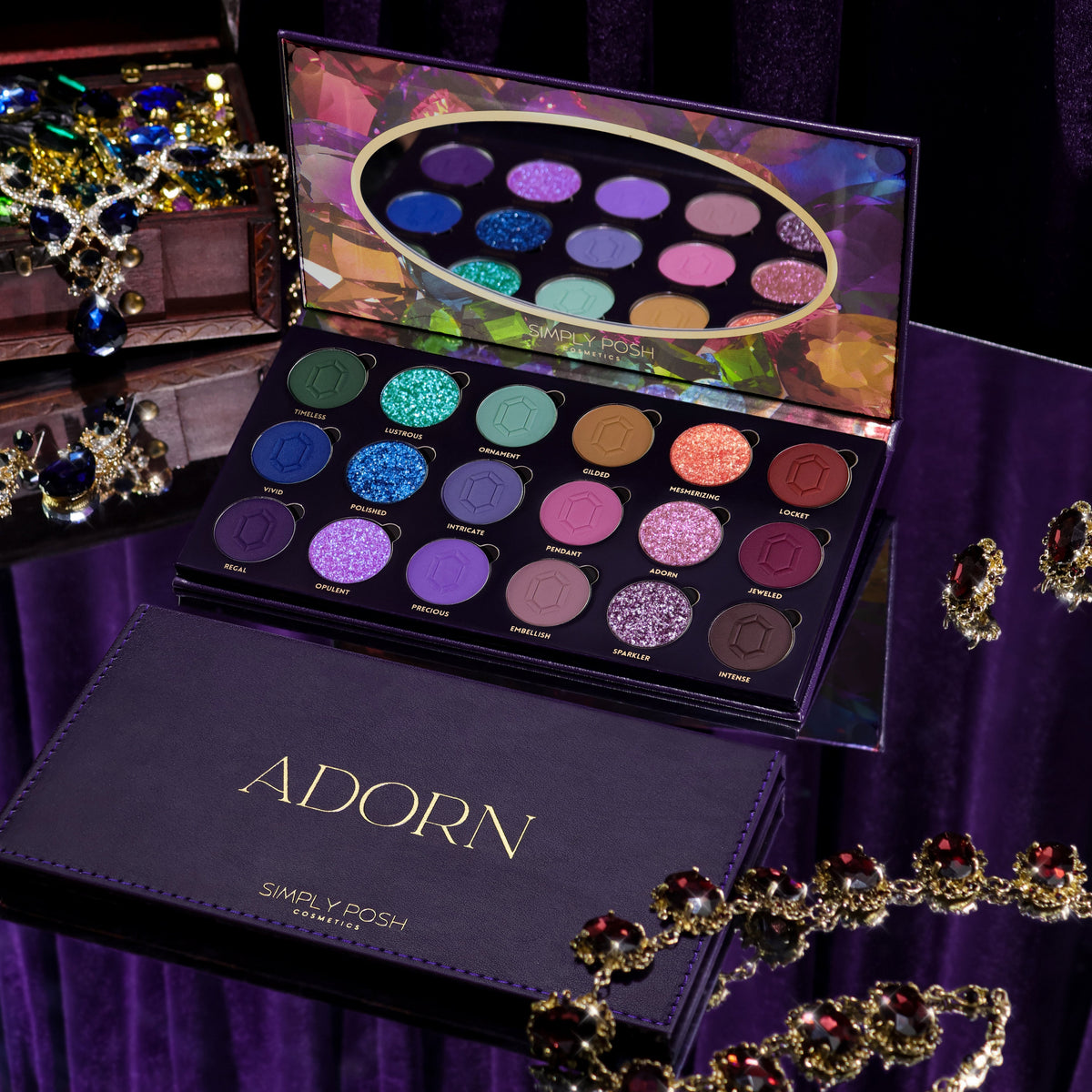 Simply Posh Adorn eyeshadow palette open beside its purple faux-leather cover with gold lettering, 18 pans of greens, blues, gold, pinks, purples, and browns