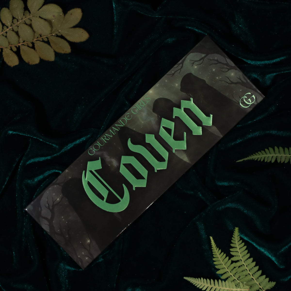 Coven palette box with green gothic lettering over shadowy figures and bare branches, on teal velvet with fern leaves