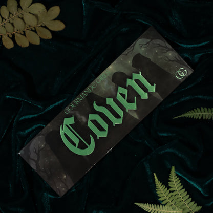 Coven palette box with green gothic lettering over shadowy figures and bare branches, on teal velvet with fern leaves