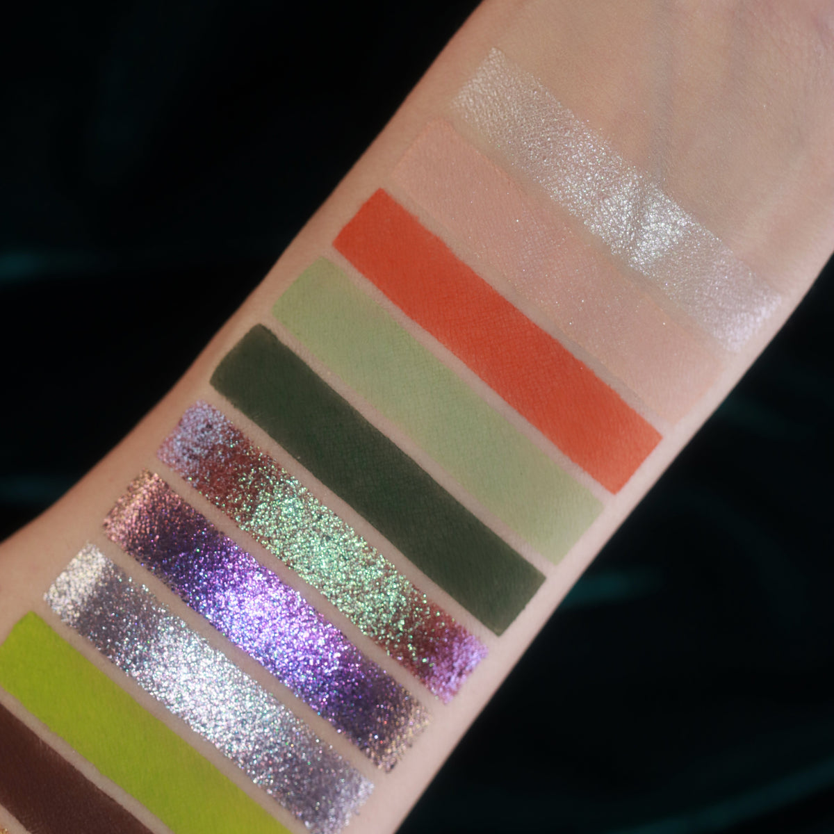 Close-up of arm swatches on light skin: pale shimmer, orange, sage and green mattes, then purple and silver sparkles