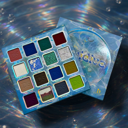 Liquidiva palette open over its lid, all 16 pans labelled with shade names, on a rippled water background