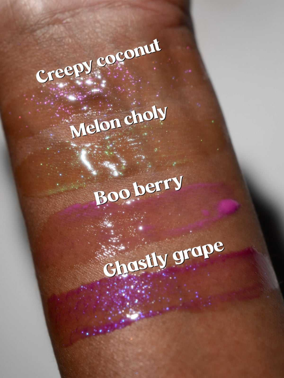 Arm swatches of the four glosses on deeper skin, labelled Creepy coconut, Melon choly, Boo berry and Ghastly grape