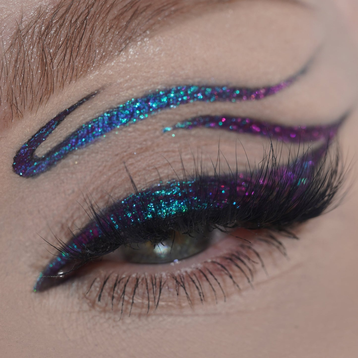 Close-up of a single eye with teal and purple multichrome graphic liner extending above the brow, paired with full black lashes.
