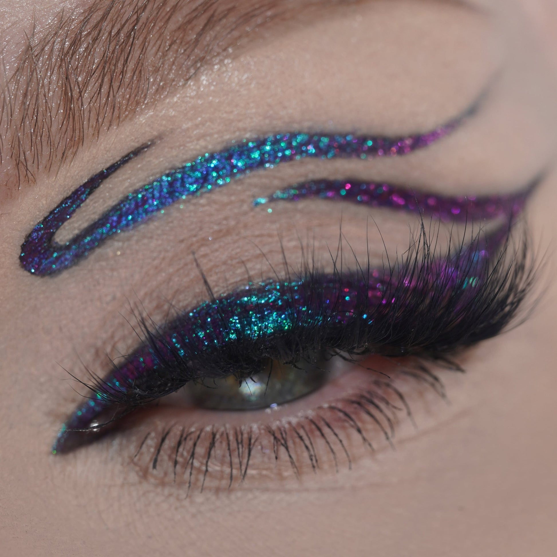 Close-up of a single eye with teal and purple multichrome graphic liner extending above the brow, paired with full black lashes.