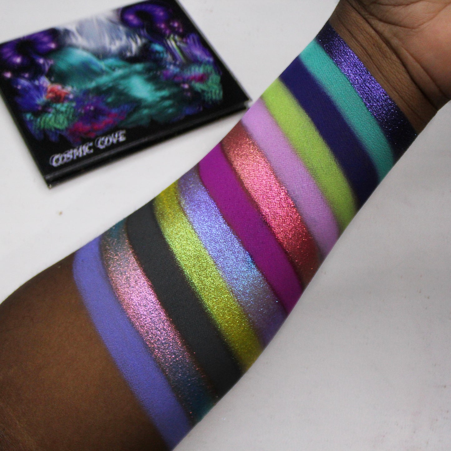 Cosmic Cove swatches on deep skin tone showing all twelve shades, with the closed palette in the corner showing the painted cover artwork