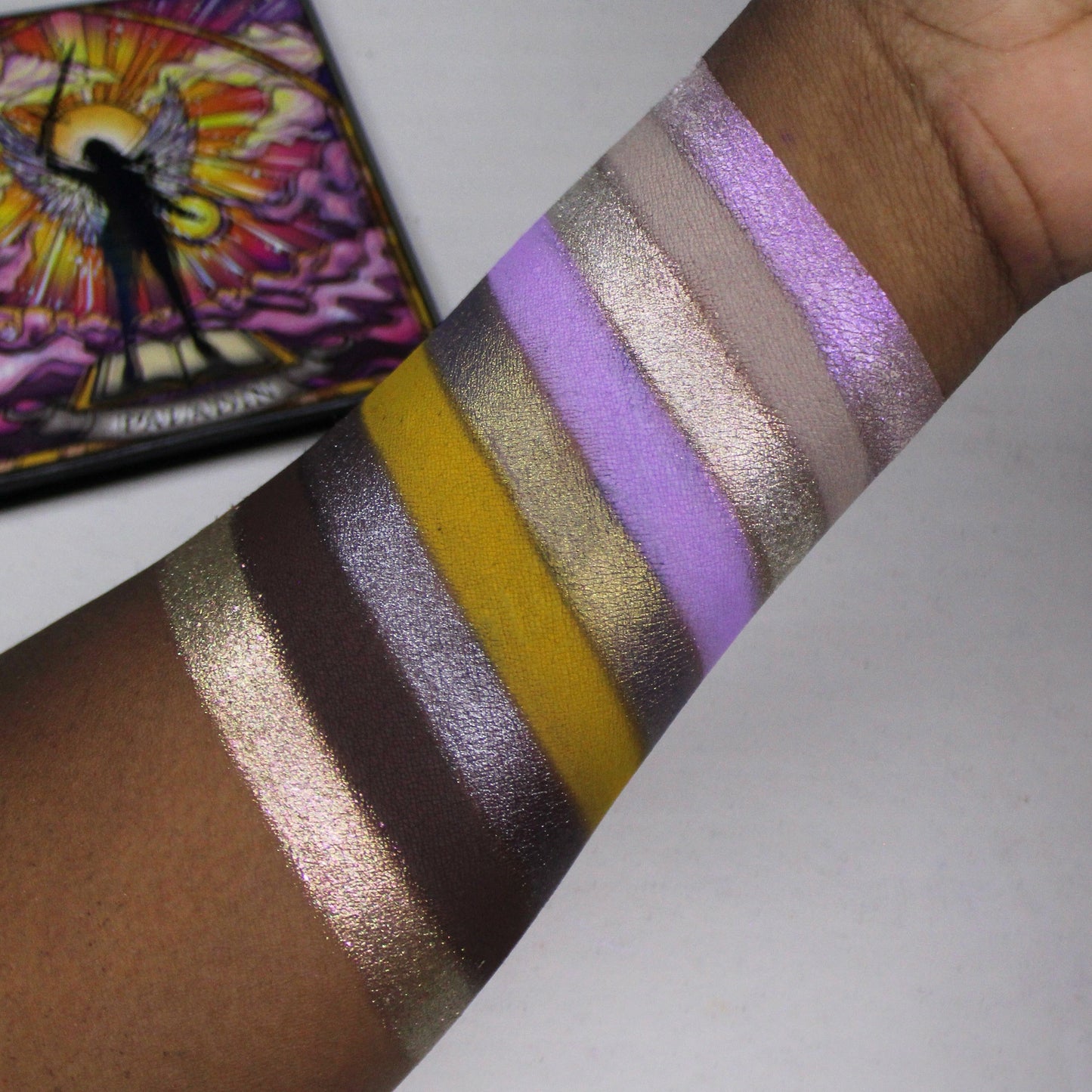 Paladin swatches on a deep skin tone arm, nine stripes running gold sparkle, deep brown, silver purple, mustard, lilac and pink shimmer, closed palette behind