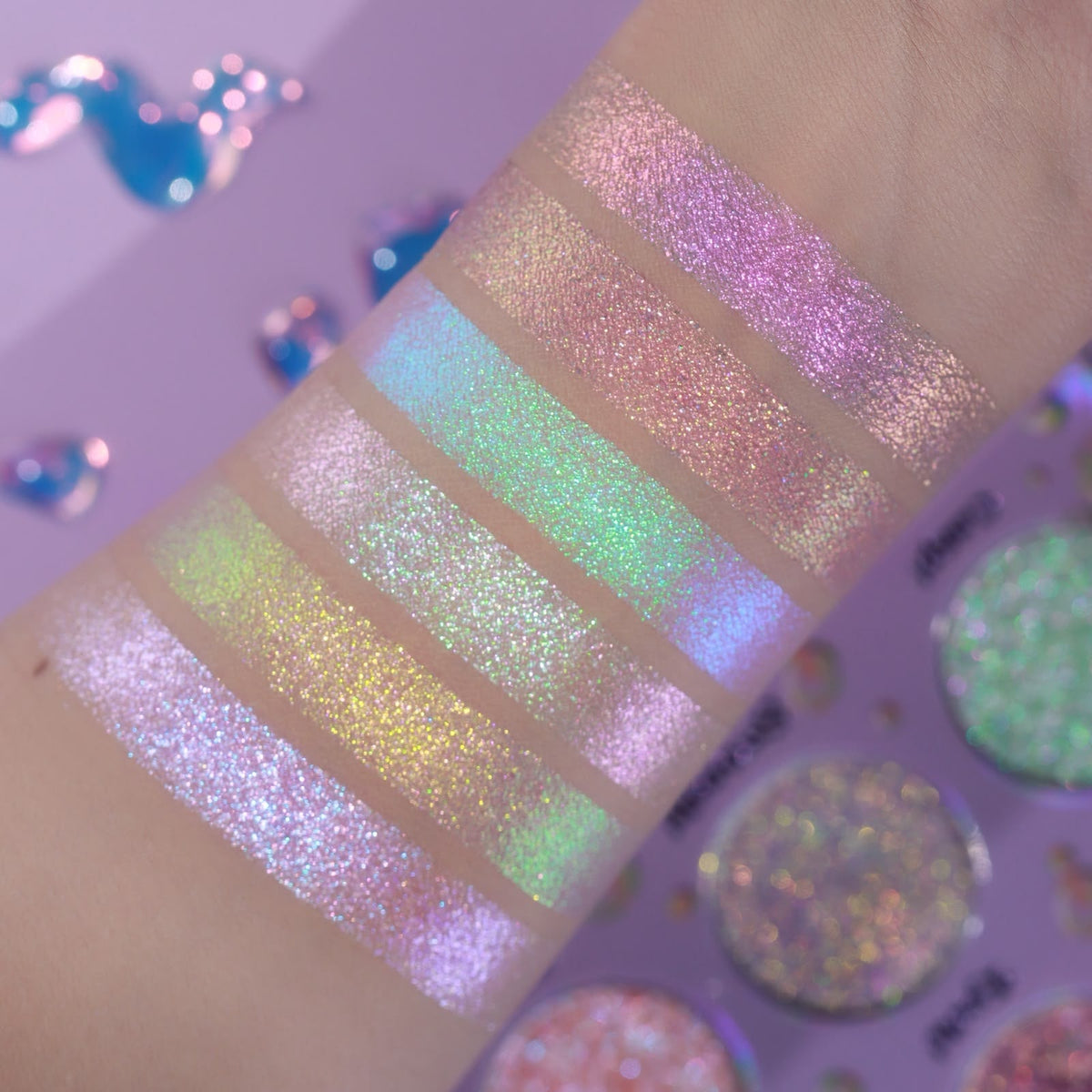 Unlabelled arm swatches on light skin against a lilac background, with three of the palette pans visible at the right.