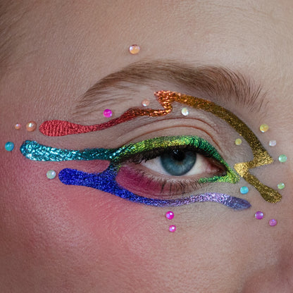 Close-up of a blue eye with a rainbow gradient drip liner from red to violet, surrounded by scattered pastel face gems.
