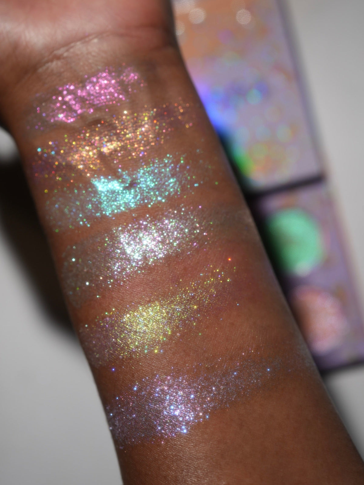 Unlabelled arm swatches on deep skin at an angle, with the open palette propped behind at the right.