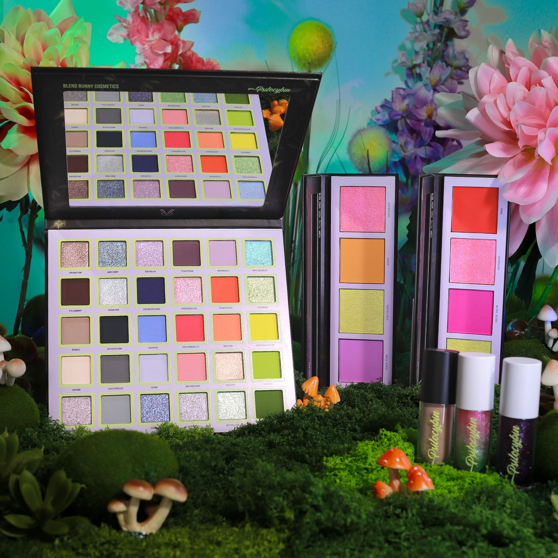 Full Psilocybin collection on moss, the open 30 shade eyeshadow palette beside two cheek palettes and three lip glosses on a floral backdrop