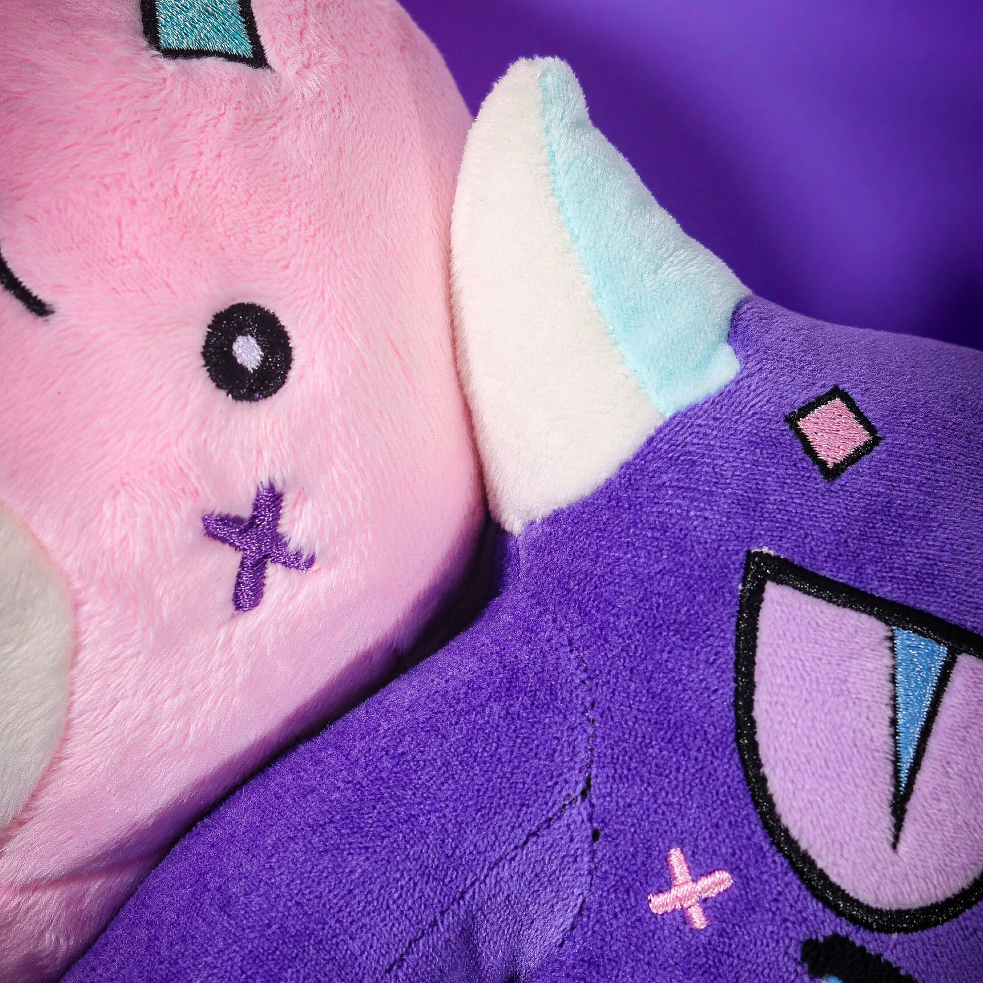 Close-up of the Gleam and Gloom plush faces side by side, showing the embroidered stitch details.